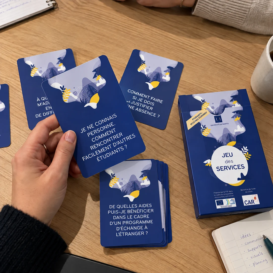 Partie du Jeu des services : une main tient la carte « Je ne connais personne. Comment rencontrer facilement d'autres étudiants ? », à côté de l'étui du jeu